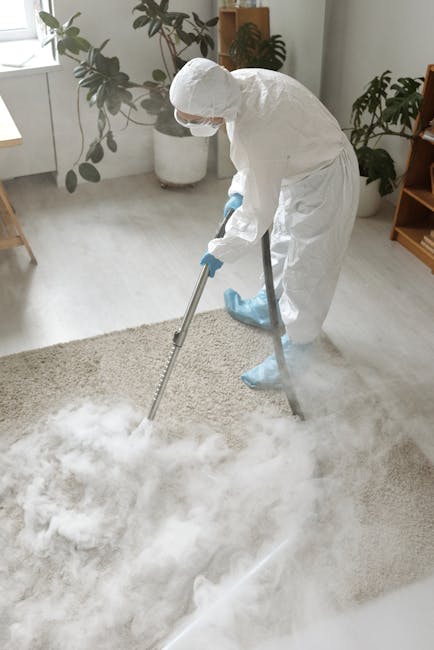 A person kneeling on a patterned carpet in a residential room, operating a yellow and black vacuum cleaner with a flexible hose. The individual is wearing a beige shirt and blue jeans, and is engaged 