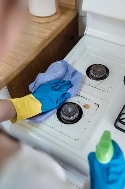 Close-up of a person's hand wearing a yellow cleaning glove, using a blue cloth to wipe a stained and greasy cooktop surface with two round burners on a white ceramic stove. The stove is situated in a